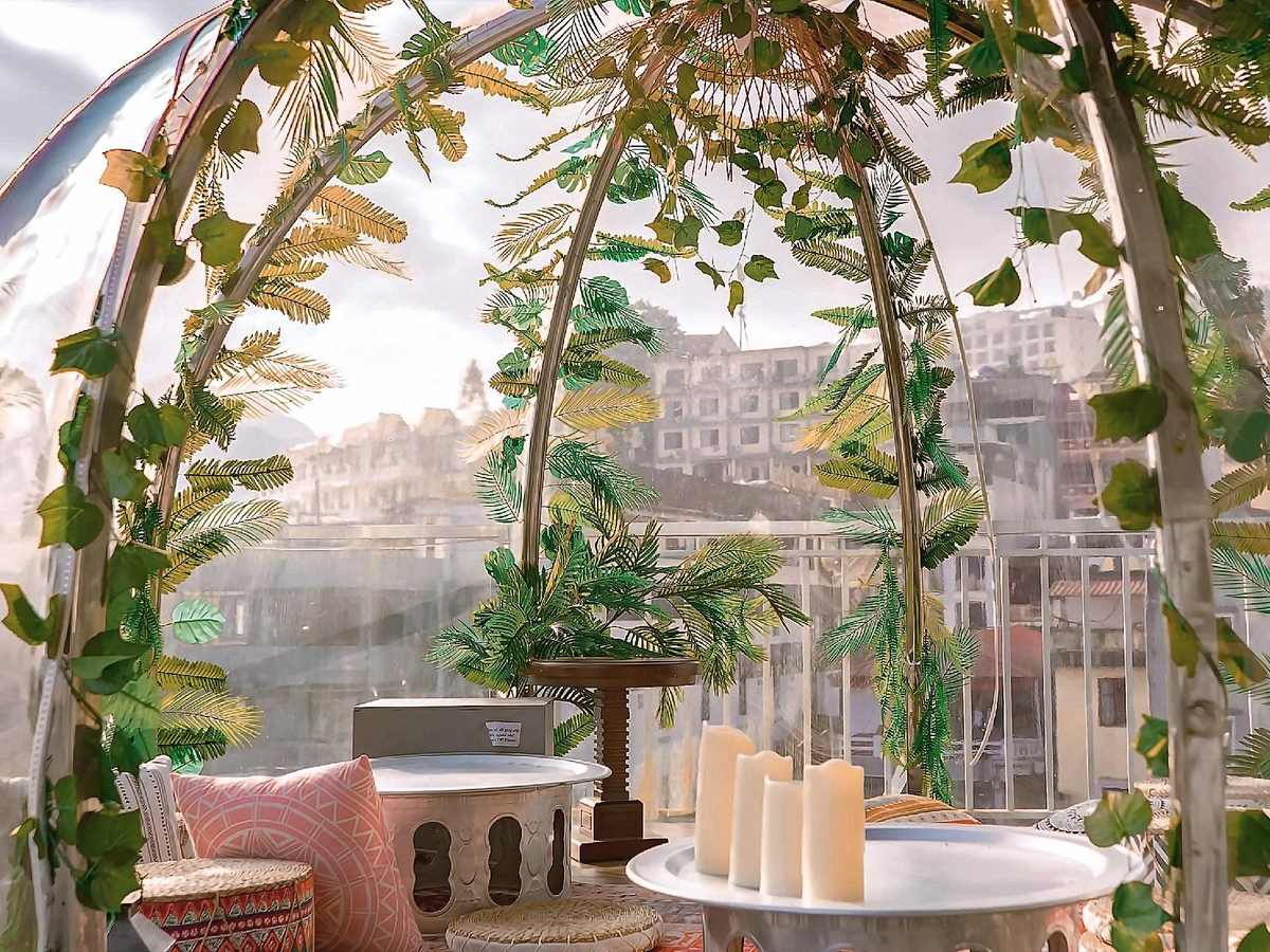 Inside a clear dome wrapped in green vines, with cushions, white candles and town buildings seen through the curved wall