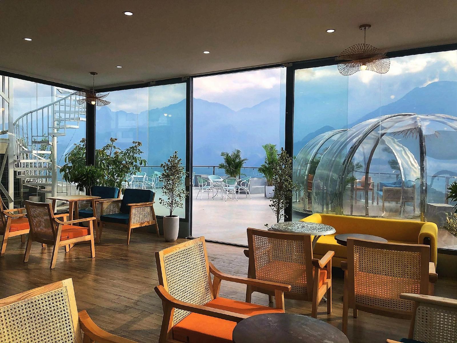 Glass-walled caf&eacute; with rattan chairs and orange cushions, a spiral staircase and a clear dome on the terrace outside, mountains beyond