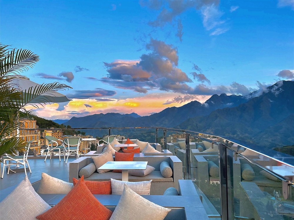 Rooftop terrace with grey sofas and orange cushions behind a glass railing, mountain ridges under a sunset sky beyond