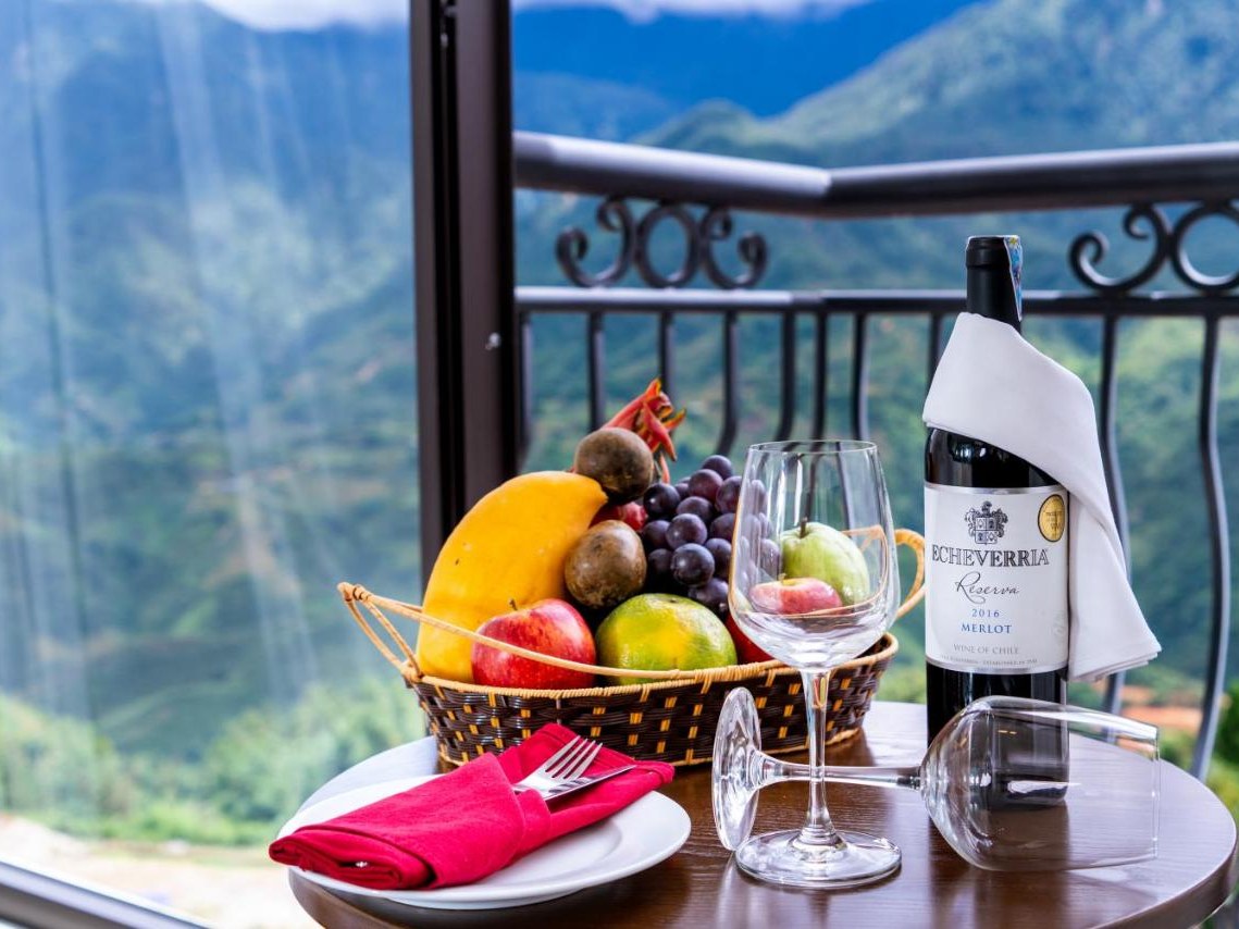 Fruit basket, a bottle of wine and glasses on a balcony table, with a green mountainside beyond the railing