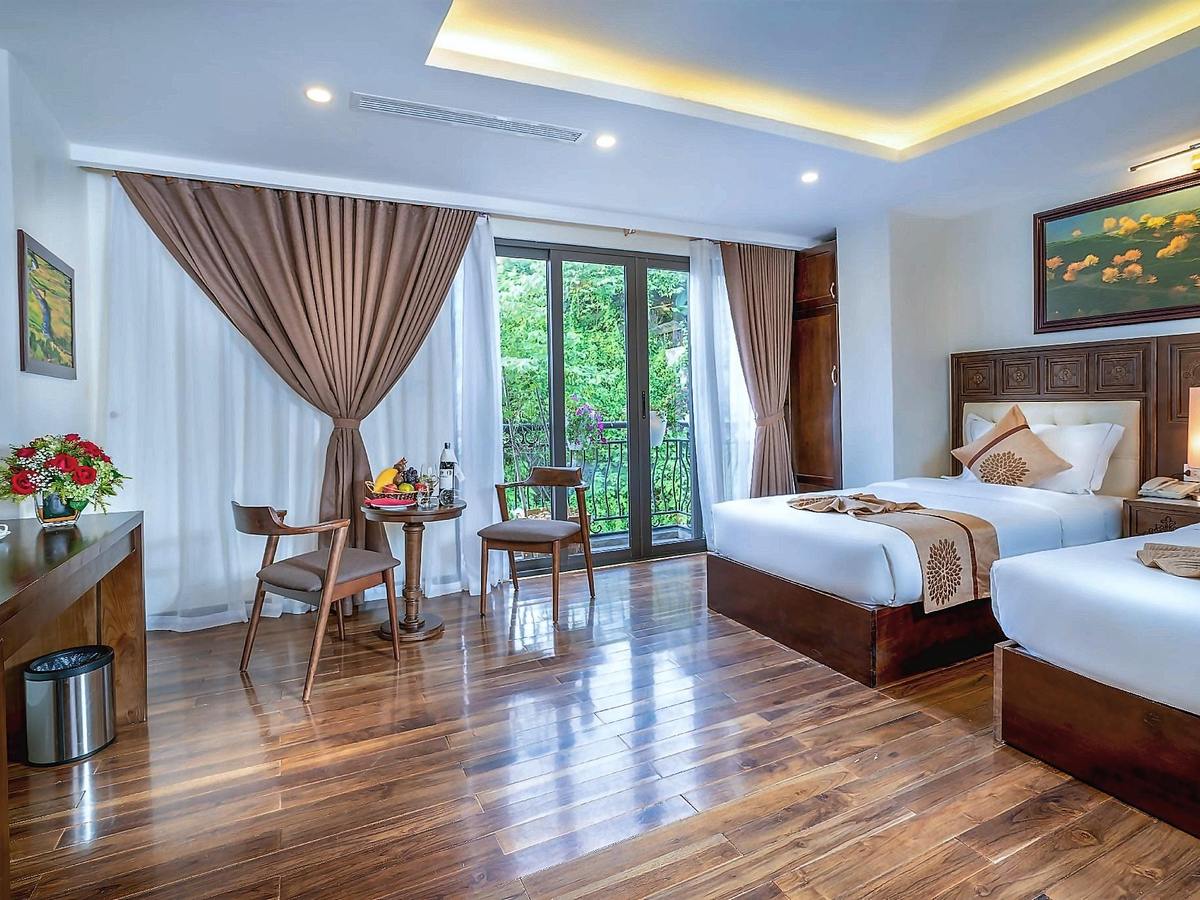 Twin room with dark wooden floor, carved headboards, beige curtains and a glass door to a small balcony facing trees