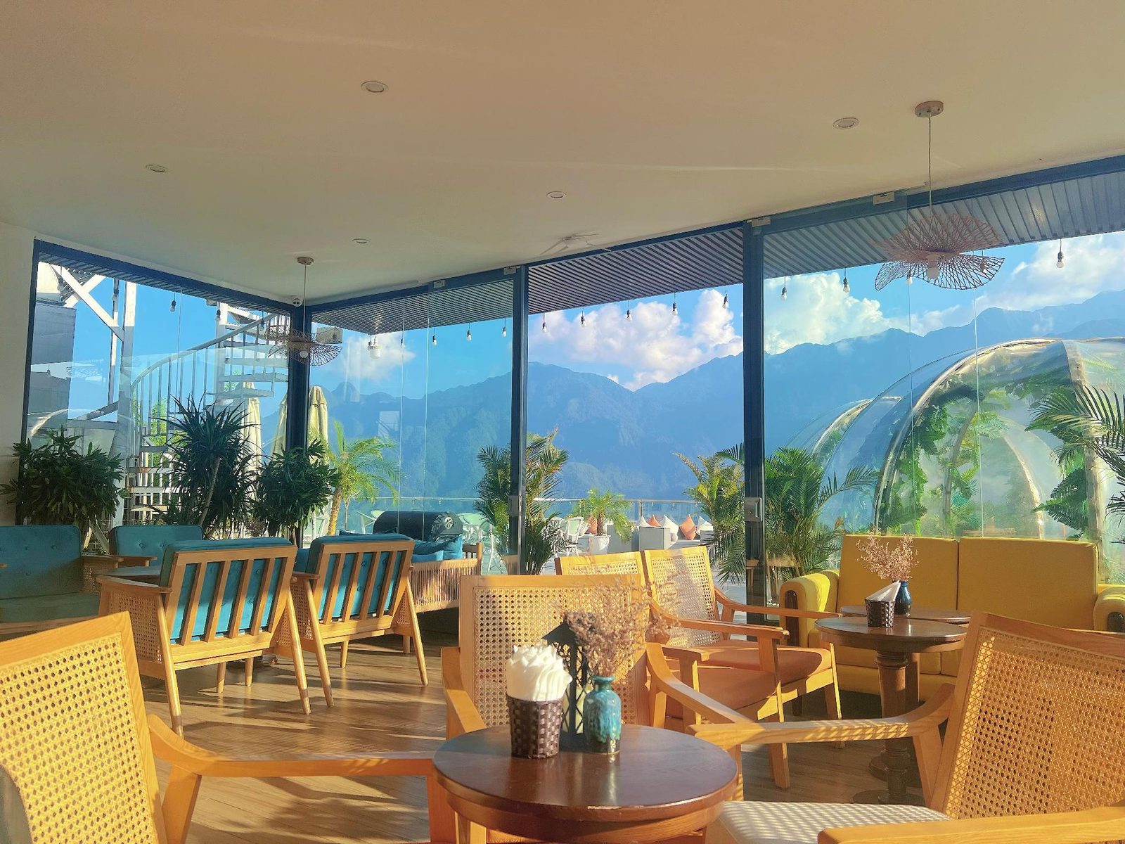 Glass-walled rooftop café with rattan armchairs, teal cushions and a yellow sofa, a clear dome outside and green mountains beyond