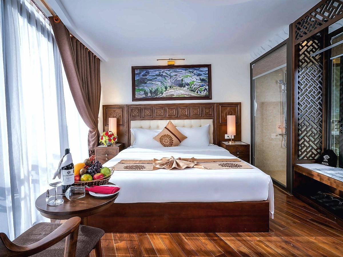 Double bed with a carved headboard, a fruit plate on a round side table and a glass-walled bathroom with a wooden screen