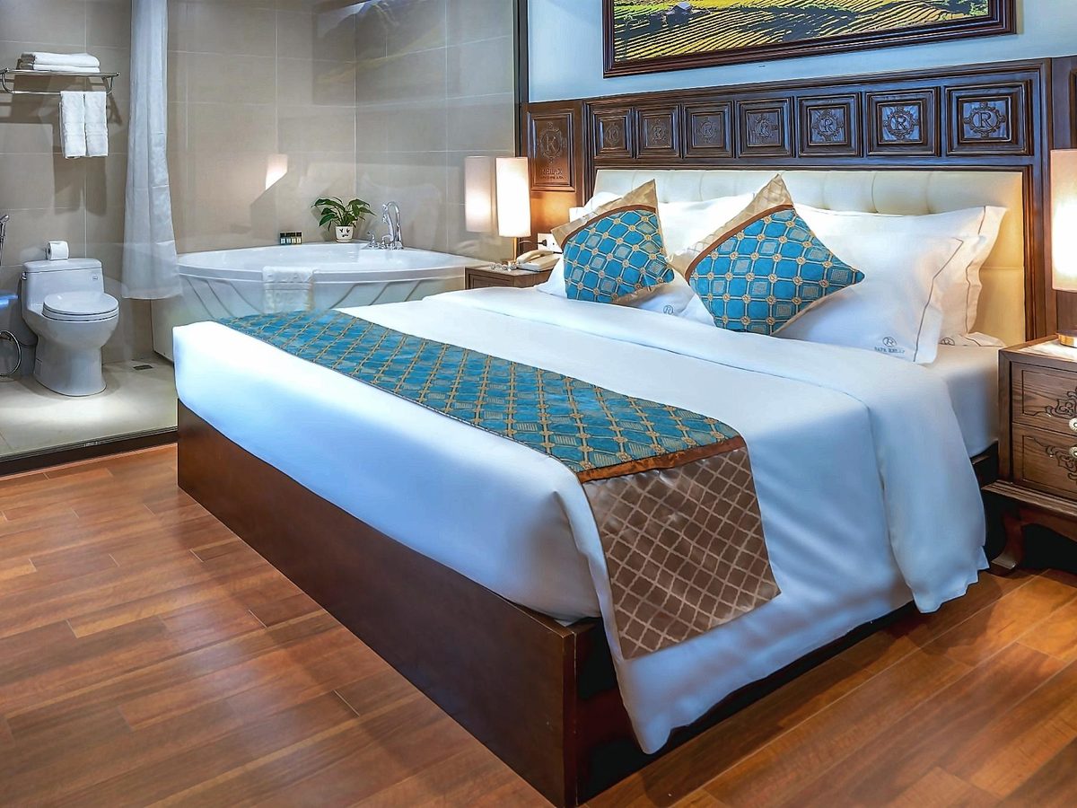 Double bed with teal cushions, a corner bathtub and a toilet visible in the bathroom area behind it